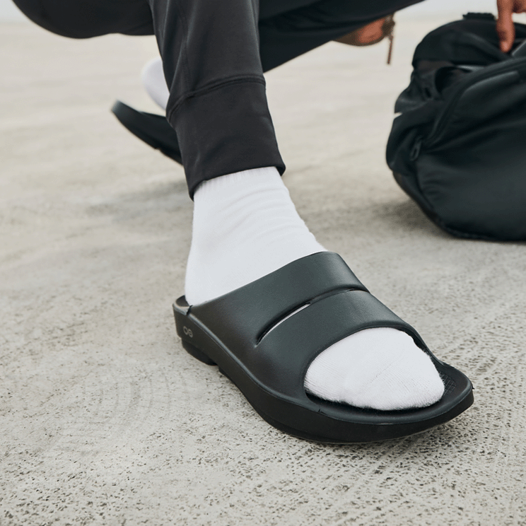 OOFOS OOahh Slide — Black, close-up on foot outside #Colour_Black